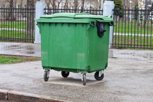 A large green plastic wheeled waste bin with a closed lid is positioned on a paved surface in front of a black metal fence, which encloses a grassy area with trees and pathways. The bin has a textured surface with slight scuff marks and is situated outdoors, likely in an urban or suburban setting. Nearby, the ground appears damp, possibly from recent rain, and the environment suggests it could be a street or communal area in a residential neighbourhood, such as Poplar. The image relates to rubbish collection and waste management services provided by House Clearance Poplar, emphasizing the importance of proper rubbish disposal in local communities.