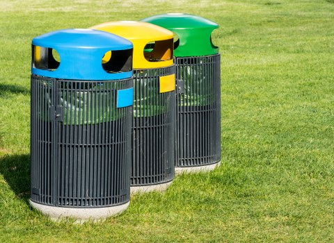 Three cylindrical outdoor rubbish bins with black plastic body and vertical slats are positioned on a well-maintained grassy area, likely on a driveway or communal space in Poplar. The bins have brightly coloured, rounded lids in yellow, blue, and green, each with a small rectangular label on the front, indicating different waste types. The yellow-lidded bin is on the left, the blue in the middle, and the green on the right. The background shows lush green grass and a slightly blurred outdoor environment, suggesting a residential or communal setting. The bins are clean and appear to be used for household waste collection, typical of rubbish removal services provided by companies like House Clearance Poplar. The lighting indicates natural daylight, and the overall scene conveys a typical refuse management setup in East London or nearby areas, emphasizing the importance of proper waste disposal and clearance.