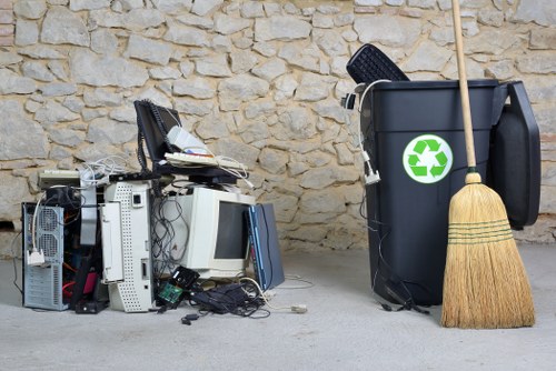 In the foreground on the left side of the image, there is a pile of discarded electronic waste, including an old computer monitor, keyboards, wires, and other hardware components, all stacked loosely on a concrete surface. To the right of this pile, a large black rubbish bin with a green recycling symbol is positioned against a textured stone wall, and a wooden broom with natural bristles leans against the bin. The scene appears to depict a collection point for waste or rubbish awaiting removal, situated in an outdoor area possibly adjacent to a residential or commercial property in Poplar. The lighting is natural, suggesting daytime, and the environment is clean but clearly used for waste collection and disposal, consistent with rubbish removal services provided by House Clearance Poplar in the local area.
