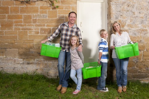 A family of four, consisting of a man, a woman, a young girl, and a boy, standing outdoors against a beige brick wall with a stone section, each holding a bright green plastic storage bin. The man, positioned on the left, is smiling and wearing a checked shirt and blue jeans. The young girl, standing in front of the man, is smiling and wearing a grey patterned sweater and blue jeans. The boy, standing to the right of the girl, is wearing a blue and white striped shirt and dark trousers, looking towards the woman. The woman, on the far right, is dressed in a white blouse and jeans, and she is also smiling while holding a green bin. They are standing on a grassy area, and the scene suggests they are preparing for rubbish or household clearance, with the bright green bins indicating rubbish collection or disposal, which is typical of services offered by House Clearance Poplar in East London. The environment appears natural and well-lit, fitting a typical domestic or community clearance context in the Poplar area.